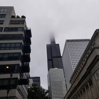 Photograph of the Willis Tower from a ground perspective. The top is poking into the clouds. 