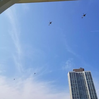 Photograph of 4 helicopters flying towards the top right of the image. In the bottom left, the top of a skyscraper is visible. 